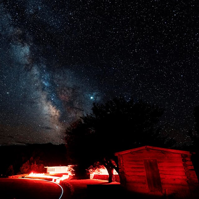 Milky Way over a red-lit Lehman Caves Visitor Center, with a historic cabin in foreground.