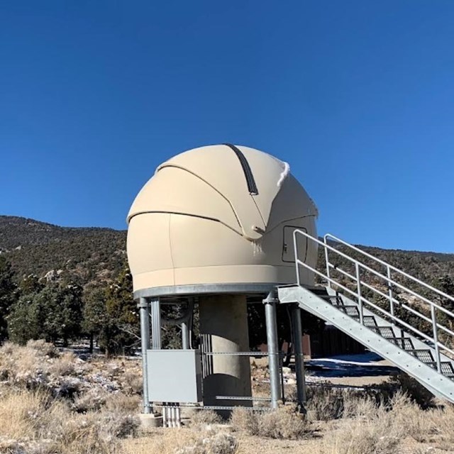 A white dome on stilts with stairs leading to it stands amongst sagebrush and before a snow mountain