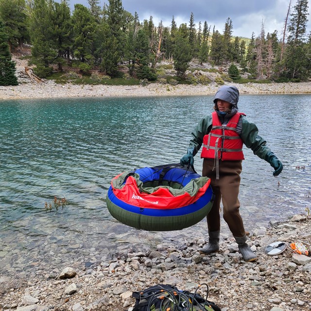 Woman in waders and lifejacket holds float tube on shore of small lake.