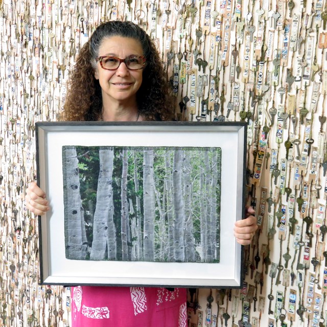 A color photo of a woman with long curly hair holding a painted quilt depicting green/white aspens
