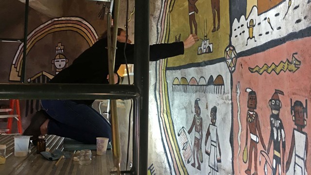 A conservator on a scaffolding renovating a mural in Desert View Watchtower.