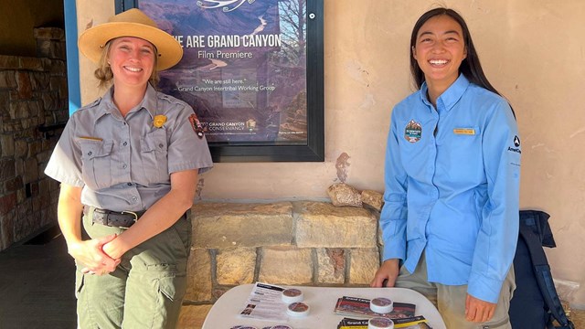 A park ranger in uniform standing with an intern wearing a blue smock by an information table.