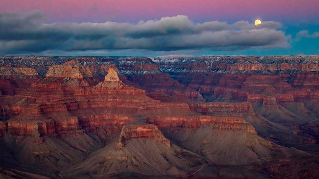 A full moon rising above a cloud bank in a sky tinted pink by sunset light above a vast canyon