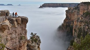 Clouds entirely filling Grand Canyon, on right, atop a massive stone pillar, two tiny people 