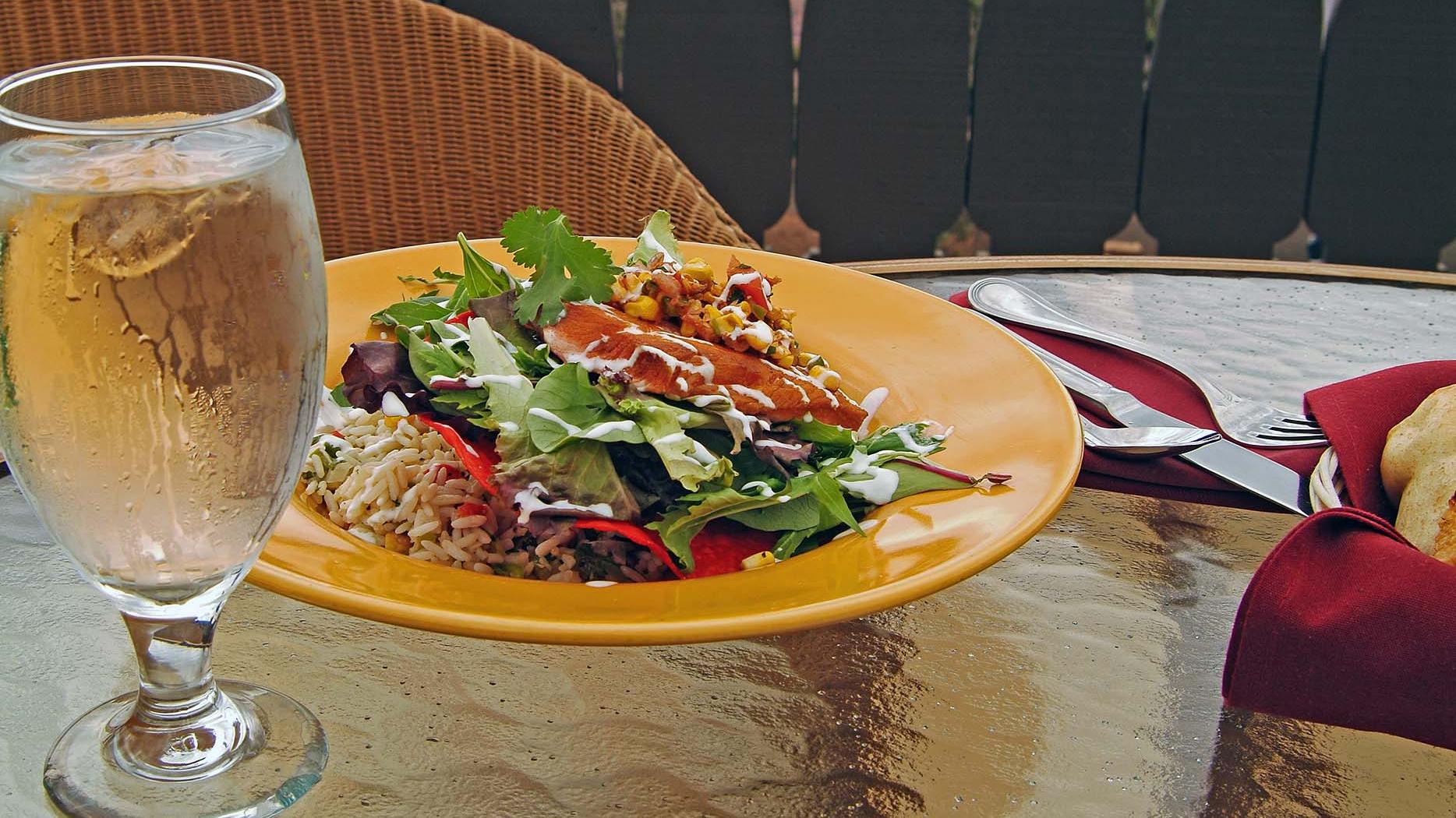 a goblet of ice water next to a fancy salad in a yellow bowl on a glass table. 