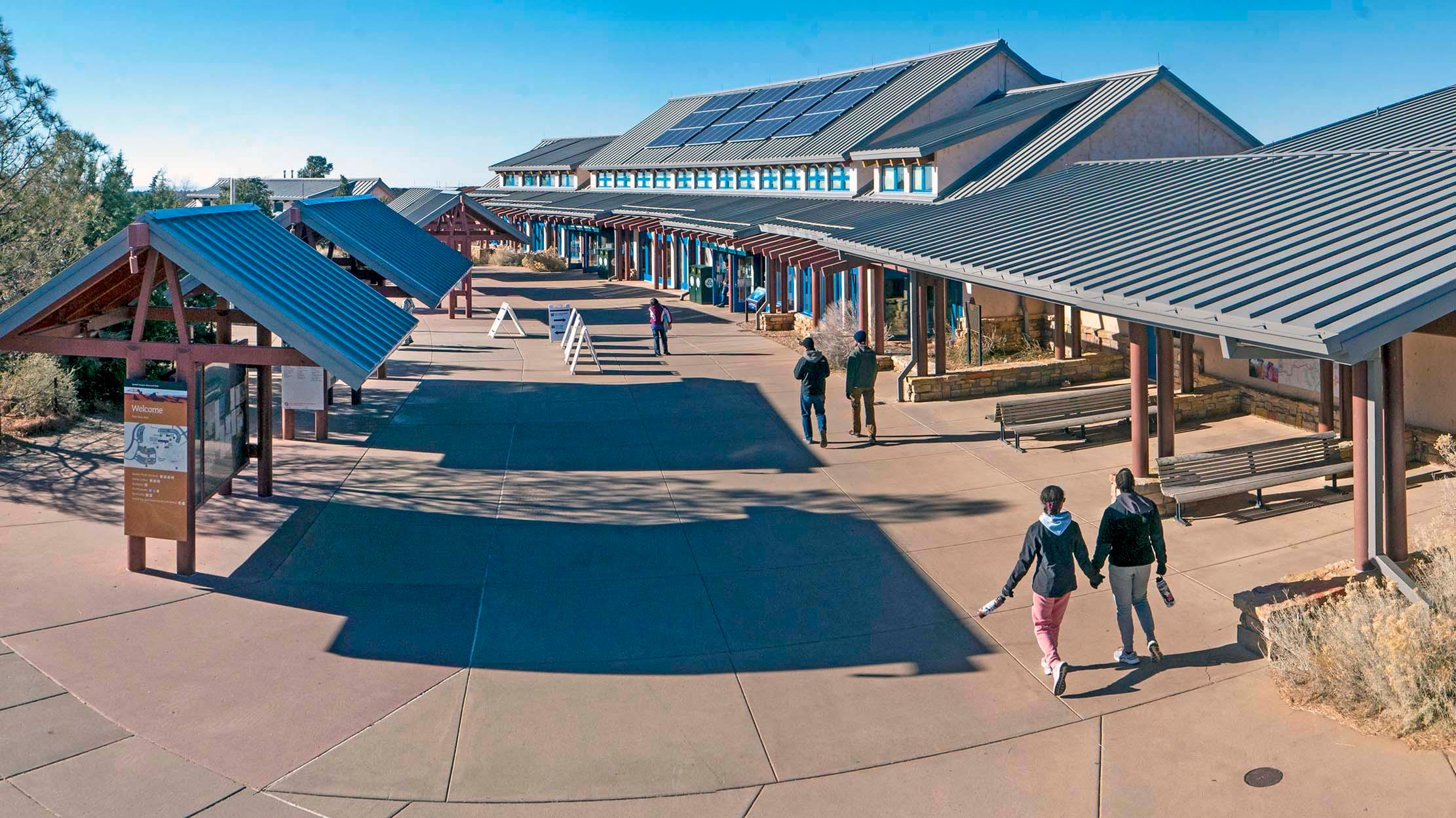 People walking across a plaza with information kiosks on the left and a long building on the right.