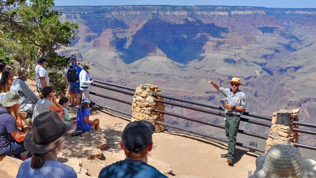 At a scenic overlook with railings, a park ranger is presenting a talk to a group of visitors.