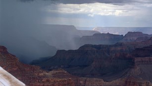 A summer storm dropping heavy rain as it casts shadows on ridgelines within a canyon landscape.