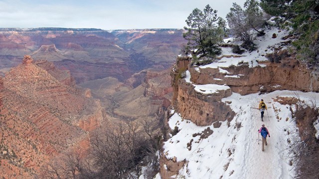 Two hikers walking on a snowy trail. Beyond them is a vast granite gorge. 