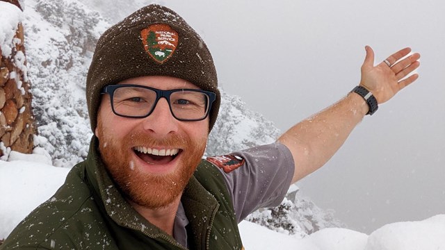 A ranger holds his hand in the air showing a snowed-in Grand Canyon landscape