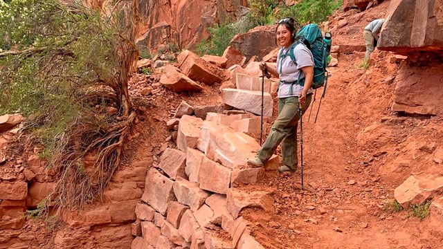 a woman holding trekking poles and wearing a backpack is standing above a rebuilt section of trail.