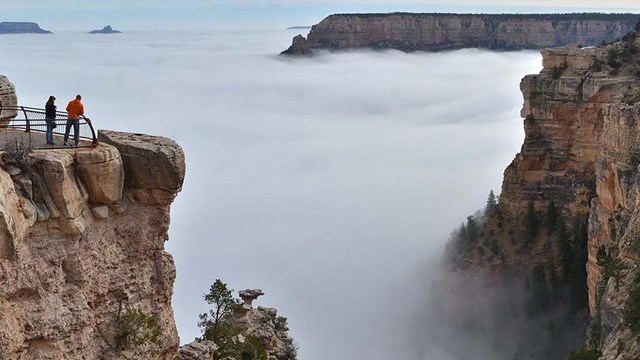 two people at a scenic overlook are looking at a vast canyon filled with a cloud inversion. 