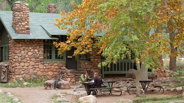 Two people sitting at a picnic table with a stone building in the background.