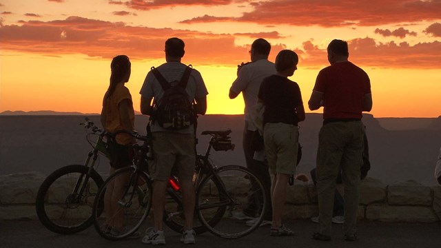 Five people in silhouette are watching a colorful sunset. Two have bicycles.  
