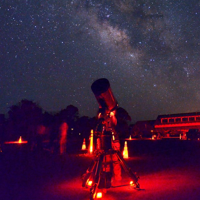 A telescope is illuminated in red light as stars that form the Milky Way shine in the background.