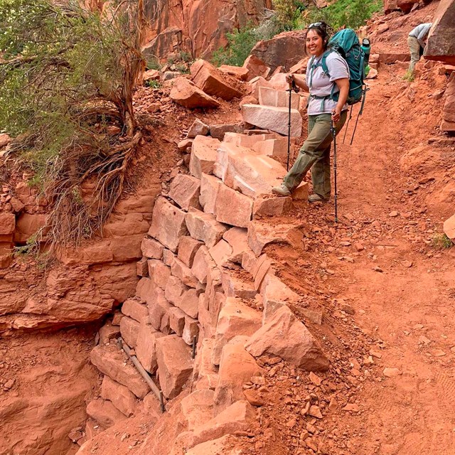 a woman holding trekking poles and wearing a backpack is standing above a rebuilt section of trail.