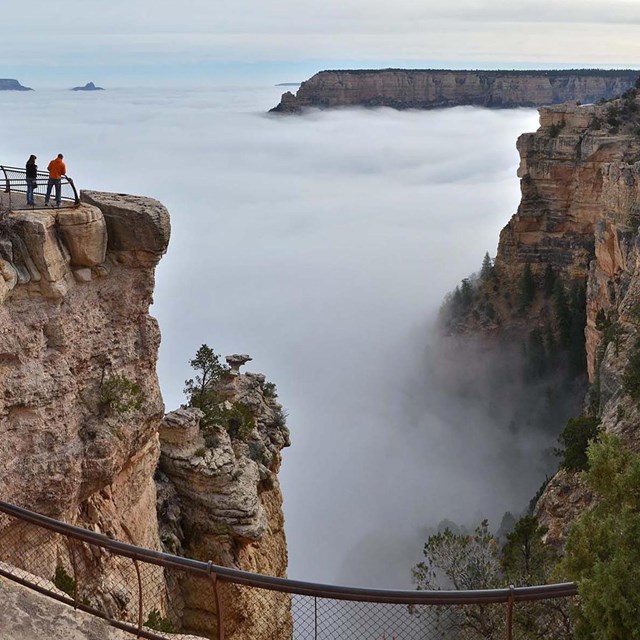 two people at a scenic overlook are looking at a vast canyon filled with a cloud inversion. 