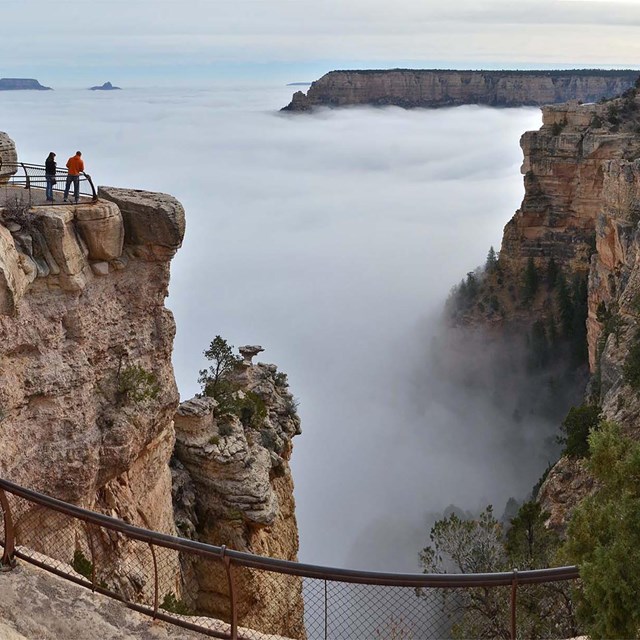 two people at a scenic overlook are looking at a vast canyon filled with a cloud inversion. 