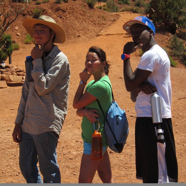 Three students make inquisitive faces at the camera while hiking on a red dusty trail.