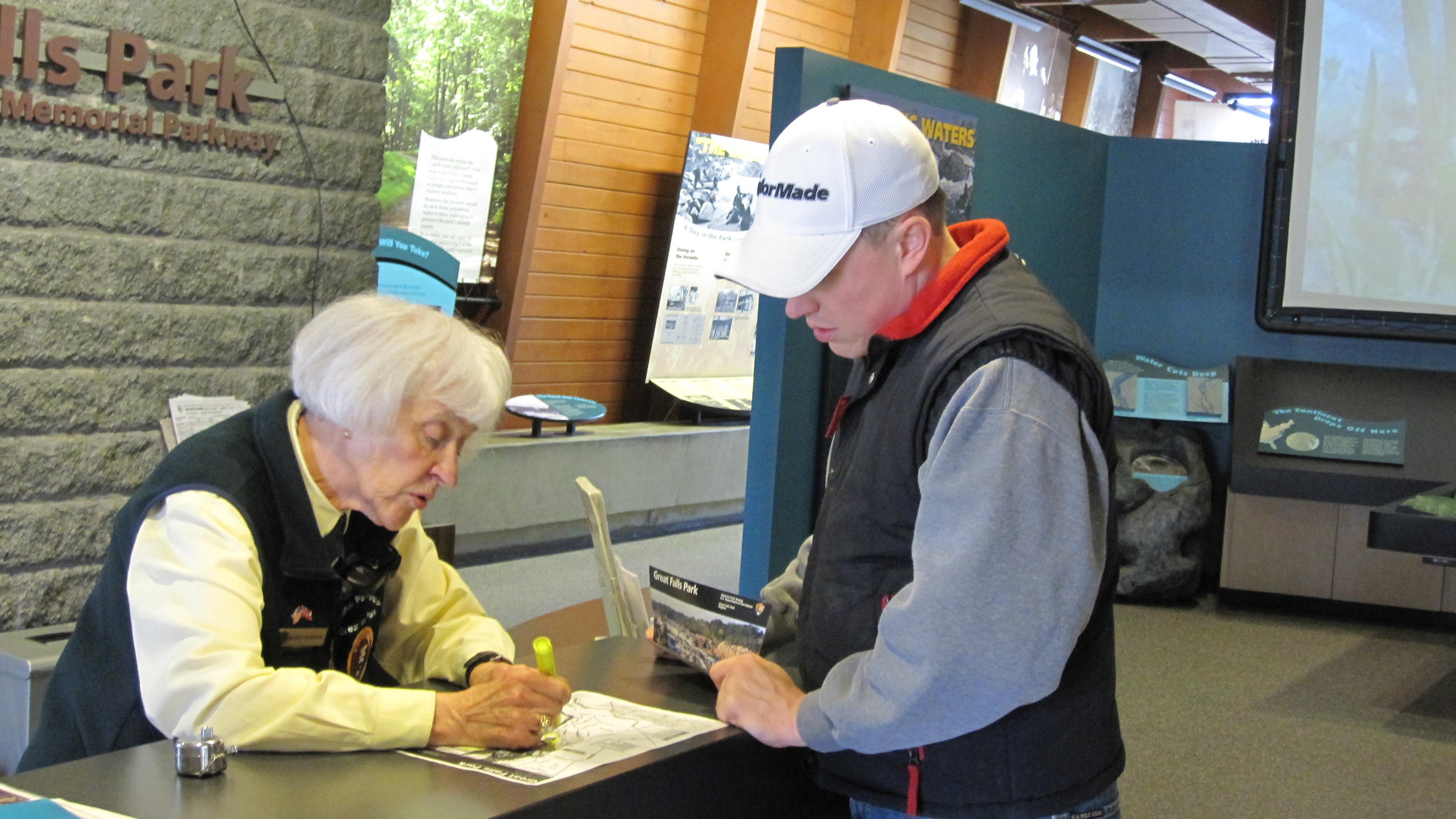 Volunteer behind desk, wearing a yellow shirt and black vest, speaks to visitor over a paper map.