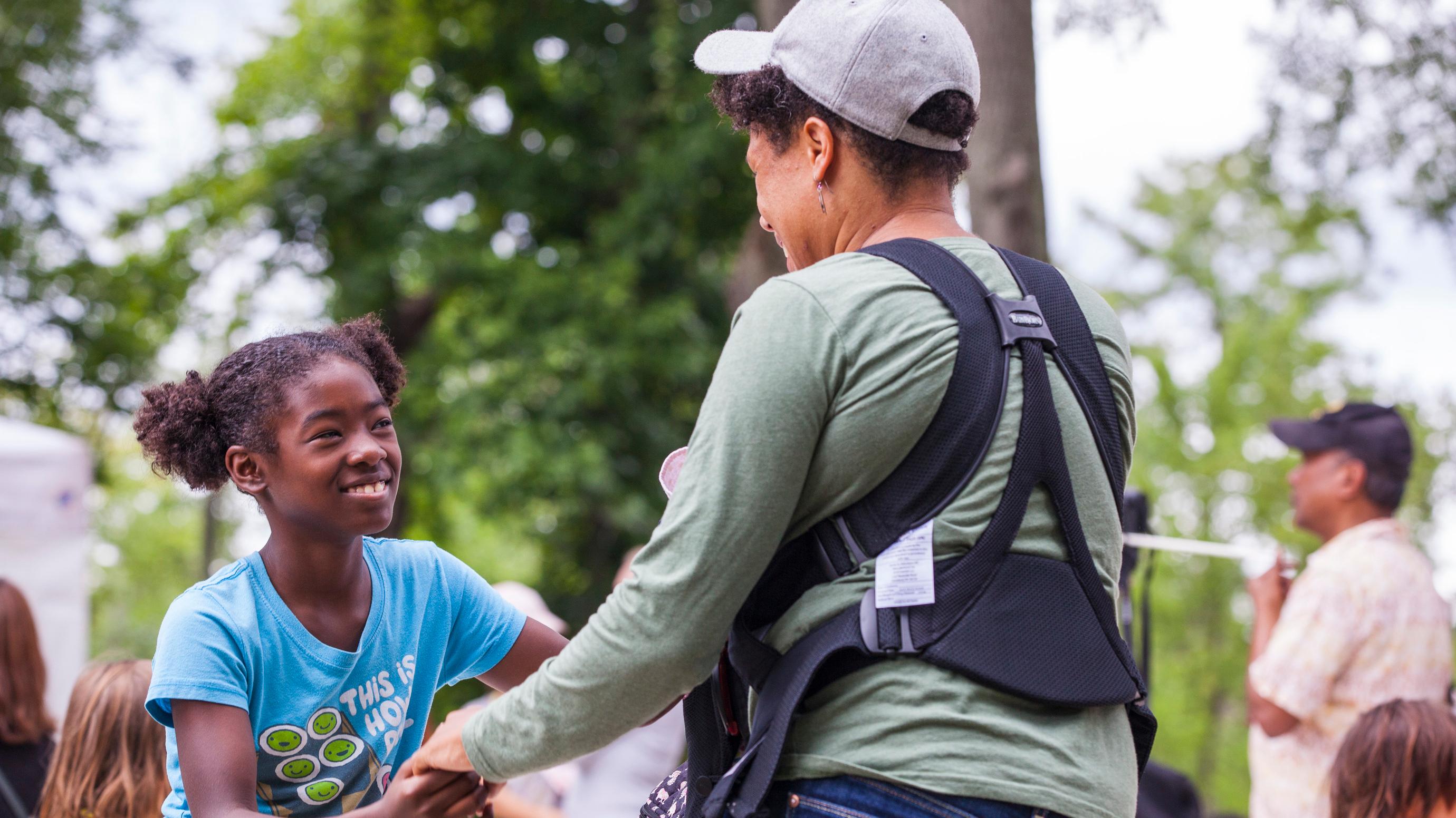 A young girl joins hands with her mother, smiling up at her, while wearing a blue t-shirt.