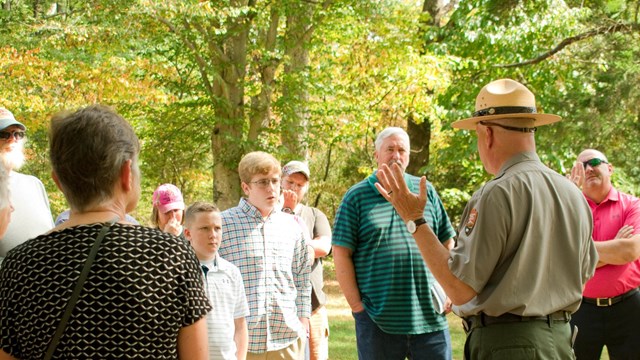 Ranger in uniform stands facing a group of visitors with his hand raised in a green forest