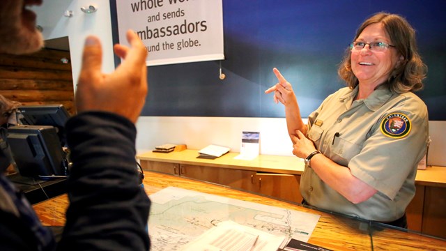 A volunteer behind a desk communicates using sign language with a visitor.