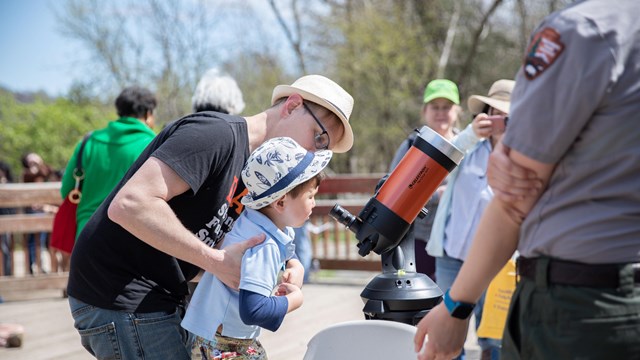 Parent lifts a child to look through a scope while a ranger watches.