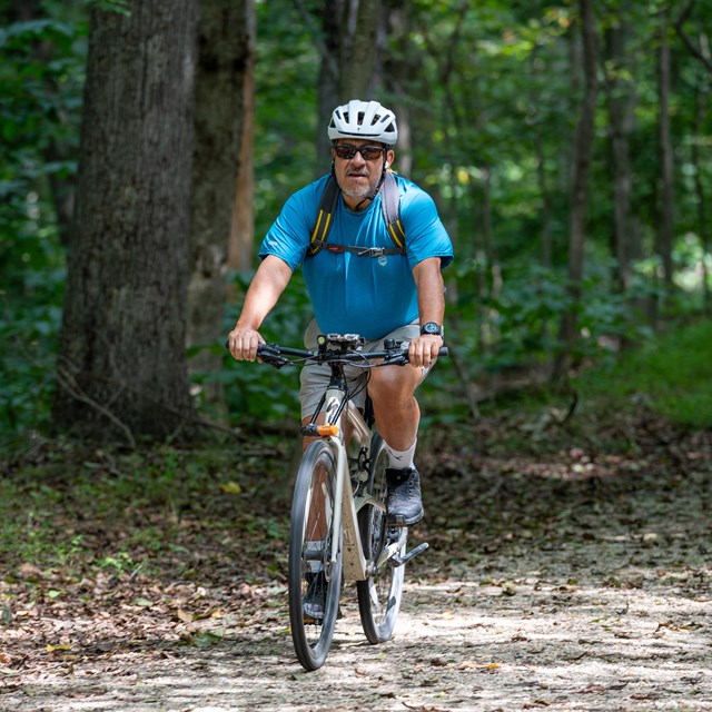 A man in a blue shirt and white bicycle helmet rides a mountain bike down a gravel trail in the fore