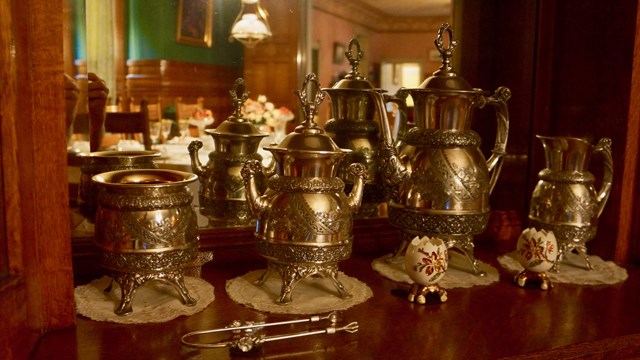 Silver tea service with four items in the set, sitting on oak sideboard with mirror behind set