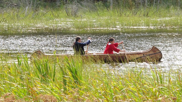 Two people wearing historic clothing, paddling a canoe on a river.
