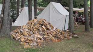 A man bent over next to a stack of wood pieces in an historic camp setting.
