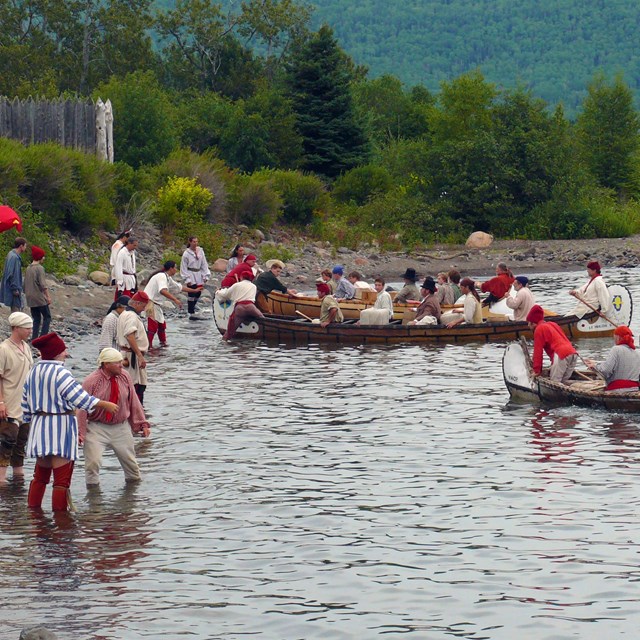 Several canoes carrying people in historic clothing land on a beach.