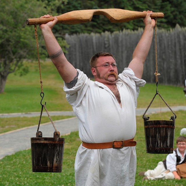 A man dressed in white with a red sash, holding up two black buckets suspended from a wood yoke.