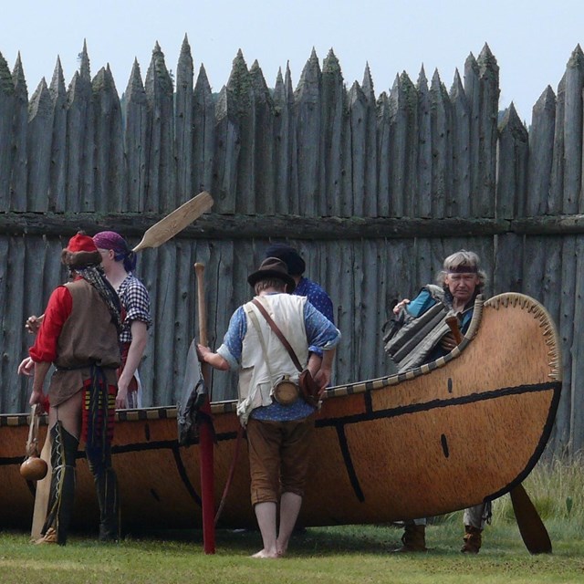 14 people in period clothing standing around a historic bark canoe in front of a wood stockade.