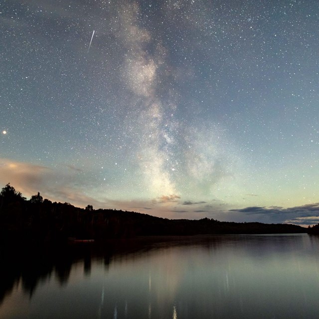 The night sky shows stars and the milky way and is reflected off a lake with a tree line on the left