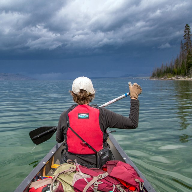 A boy in a red lifejacket paddles a canoe on a lake.