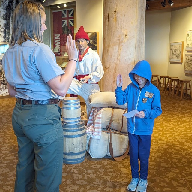 Uniformed NPS ranger with right hand raised facing a child in blue clothes with right hand raised.