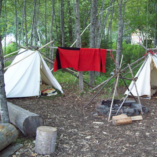 Historic white canvas voyageur tents among trees with a red blanket airing on a line.