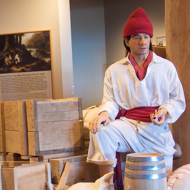 A seated mannequin dressed as a voyageur with items he is portaging.