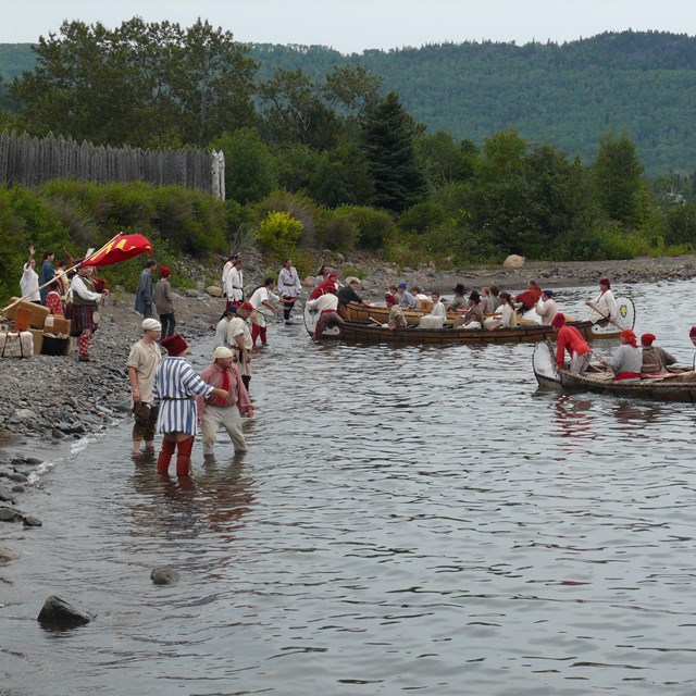 A brigade (several) canoes with people at the edge of a lake.