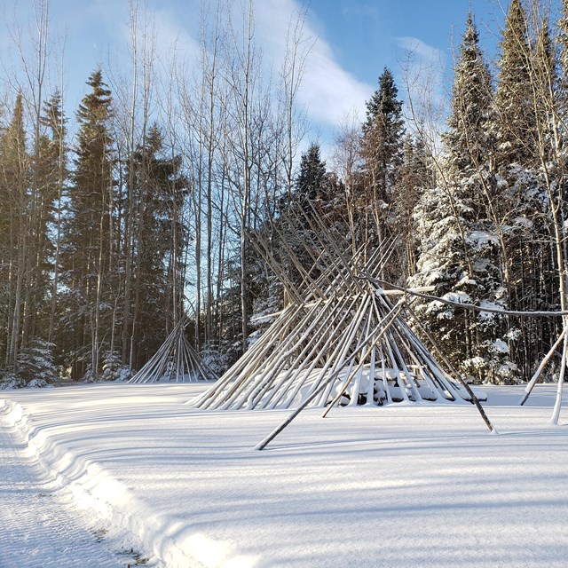 Structures made of leaning tree poles in a snowy landscape.