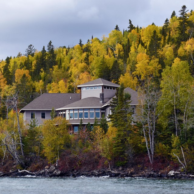 A building with a bank of windows at the base of a hill with autumn colored trees.