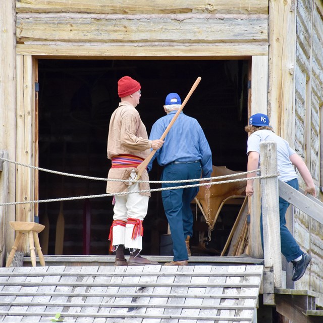 A person in historic clothing guides visitors at the entrance of a wood building.