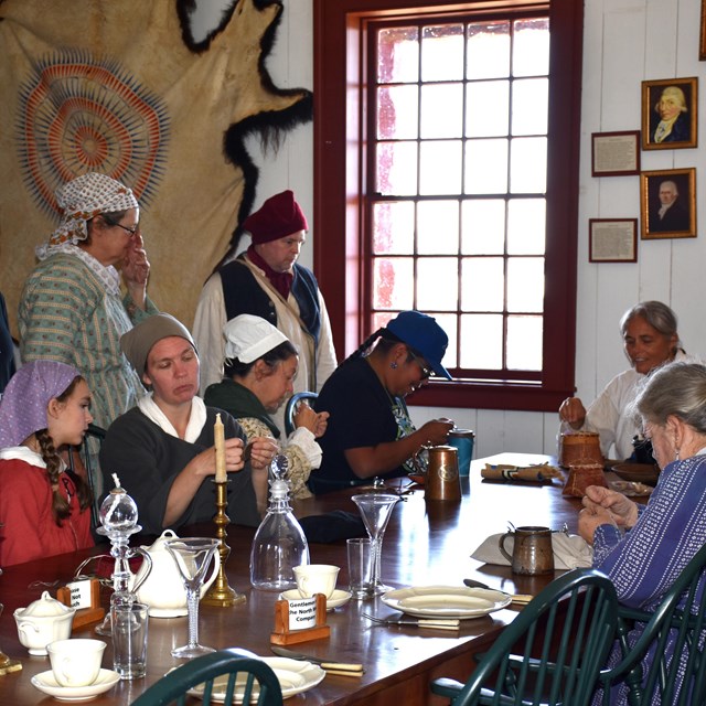 A group of people in historic clothing, sewing around a table.