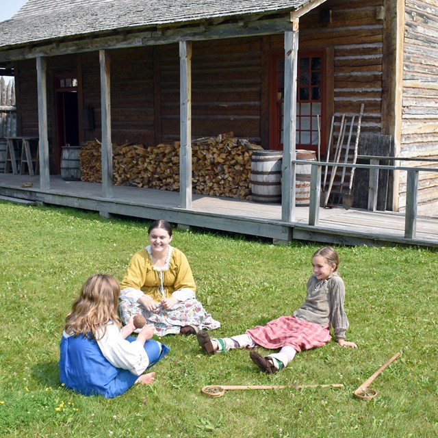 Three people in historic clothing, sitting on a lawn in front of a building.