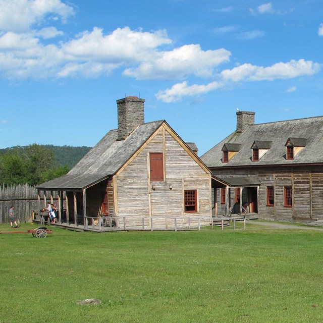 Reconstructed historic wood buildings set on a green lawn, fenced by a palisade, in front of a bay.