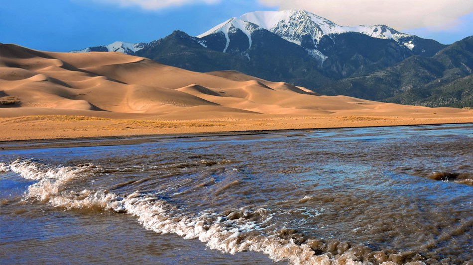 A creek with a wave at the base of dunes and snow-capped mountain