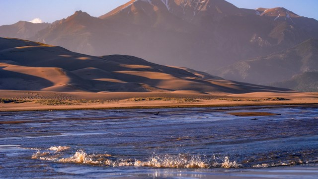 A shallow creek with a wave in front of dunes and a mountain