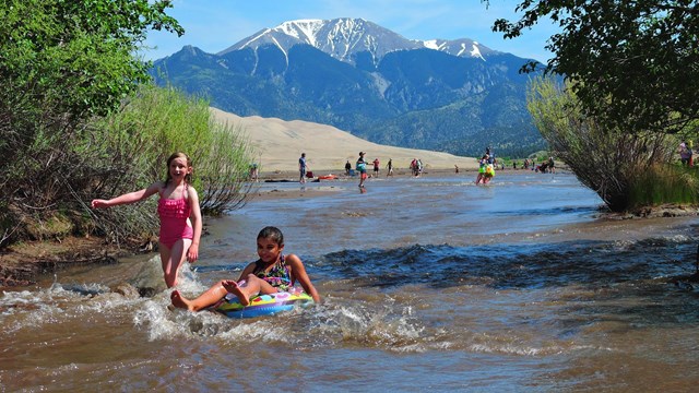 Two girls float and run on a creek with a wave, with green trees on the sides of the creek